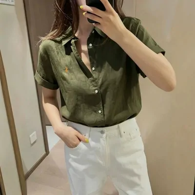 A woman in an olive short-sleeve shirt and white jeans taking a mirror photo